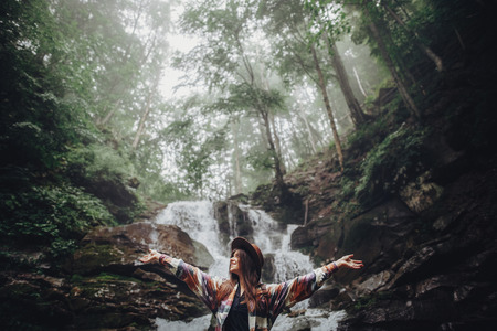 happy stylish traveler girl in hat relaxing at waterfall, atmospheric moment of success. hipster...