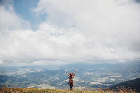 wanderlust and travel concept. girl traveler in hat with backpack raising hands up in mountains. stylish hipster woman on top of mountain in clouds. space for text. atmospheric momentの写真素材