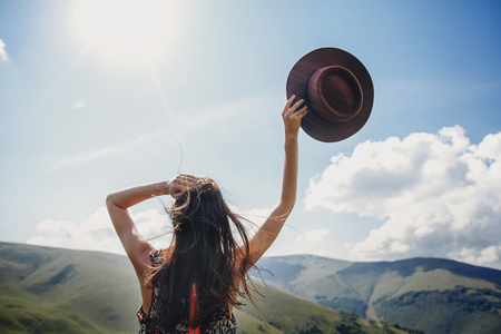 stylish traveler woman looking at mountains. hipster girl on top of mountain, raising hands and holding hat in sky. space for text. atmospheric epic moment. wanderlust and travel conceptの写真素材