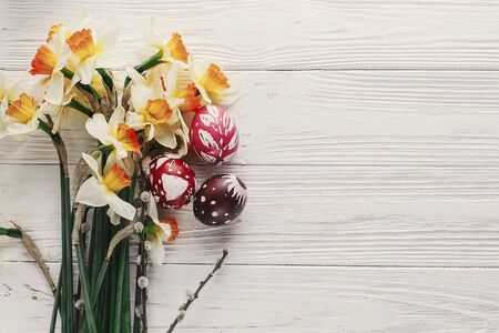 modern easter flat lay. stylish colorful easter eggs with spring flowers daffodils on rustic white wooden background top view. space for text. stylish painted eggs. greeting cardの写真素材