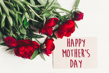 happy mother's day text sign on craft paper card beautiful red peonies on white wooden rustic...