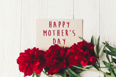 happy mother's day text sign on craft paper card beautiful red peonies on white wooden rustic background, flat lay. modern greeting card. mothers day. stylish blooming flowersの写真素材