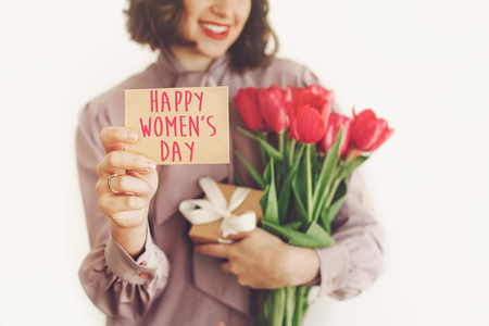 happy women's day text on greeting card. happy stylish girl holding card and pink tulips and gift, smiling on white background. happy womens day concept. young woman with flowers.の写真素材