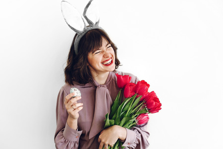 beautiful stylish girl smiling with bunny ears and pink tulips holding easter egg on white background isolated. easter hunt concept. happy woman holding painted egg. seasons greetingsの写真素材