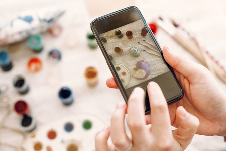 Blogging photography concept. hands holding phone and taking picture of easter flat lay with colorful eggs and paint on table. happy easter conceptの写真素材