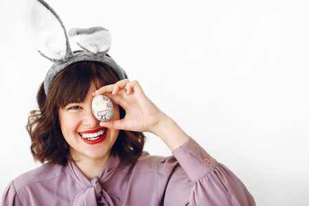 happy Easter. beautiful stylish girl in bunny ears holding colored easter egg on white background isolated. funny easter hunt concept. happy woman smiling with egg at face. fun momentsの写真素材