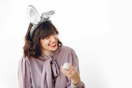 beautiful stylish girl in bunny ears holding easter egg with funny emotions and smiling on white background isolated with space for text. funny easter hunt concept. happy womanの写真素材