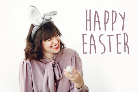Happy Easter text. season's greetings card. Beautiful happy girl in bunny ears holding colored...