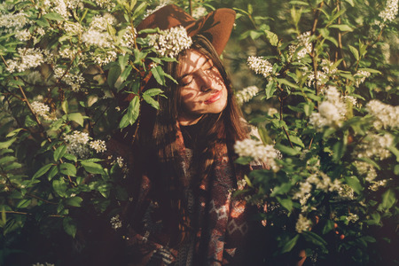 beautiful hipster girl in hat posing in green leaves with flowers in evening sunlight among shadows.の写真素材