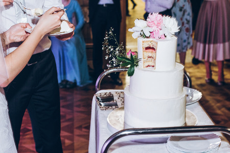bride and groom cutting delicious wedding cake at reception in restaurant. luxury wedding  couple tasting stylish white cake with peony flowers at wedding celebrationの写真素材