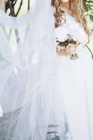 stylish bride with peony bouquet and long veil, posing in sunny garden on wedding day. luxury wedding girl, happy newlywed in silk gown. romantic momentsの写真素材
