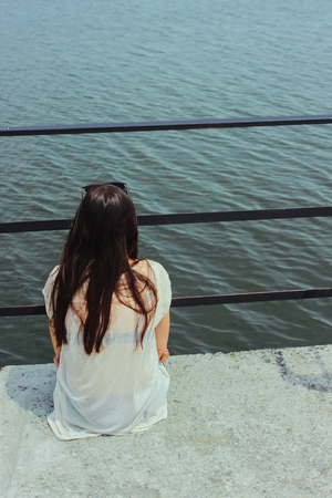 beautiful brunette girl sitting alone on a pier and looking at water in summer vacationの写真素材