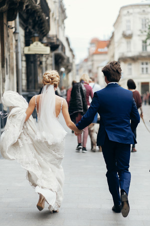 stylish bride and groom walking in city street. happy luxury wedding couple holding hands in light and running. romantic sensual moment.  back view at man and womanの写真素材