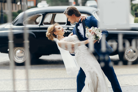 luxury wedding couple dancing at old car in light. stylish bride and groom hugging and embracing in city street. romantic sensual moment. woman looking at manの写真素材