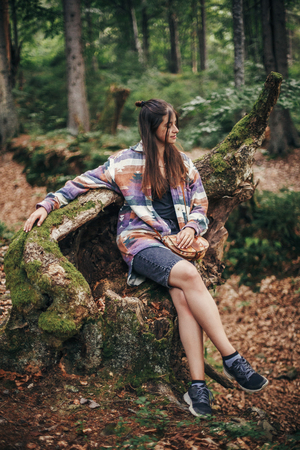 stylish traveler girl sitting on old stump with moss in sunny forest and mountains. travel and wanderlust concept. space for text. happy hipster woman relaxing in woodsの写真素材