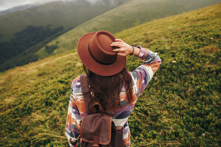 stylish traveler girl in hat with backpack looking at sunny mountains in clouds. summer vacation. travel and wanderlust concept. space for text. back view. amazing atmospheric momentの写真素材