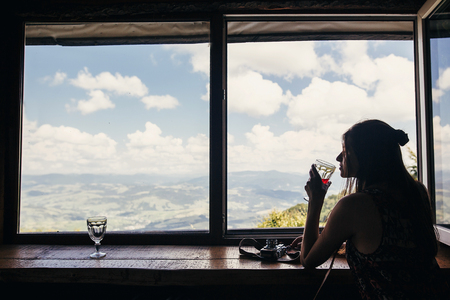 stylish girl traveler drinking at window in cottage with view on mountains and sky. happy hipster woman holding glass and relaxing. summer vacation concept. travel and wanderlust.の写真素材