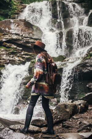 stylish hipster girl in hat with backpack looking at waterfall in forest in mountains. traveler woman exploring woods. travel and wanderlust concept. atmospheric momentの写真素材