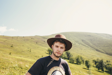 stylish traveler man in hat with herb in mouth, walking on top of sunny mountains. travel and wanderlust concept. space for text. happy hipster traveling and camping. summer vacationの写真素材