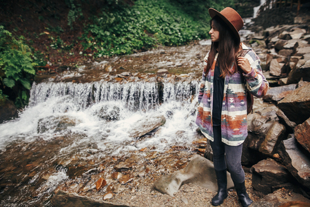 stylish hipster girl in hat with backpack standing at waterfall in forest in mountains. traveler woman exploring woods. travel and wanderlust concept. atmospheric momentの写真素材