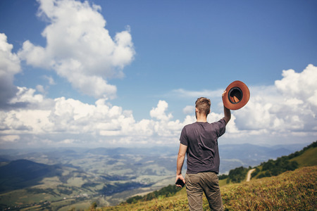 stylish traveler man with hat holding phone, walking on top of sunny mountains and sky. travel and wanderlust concept. space for text. happy hipster traveling. summer vacationの写真素材