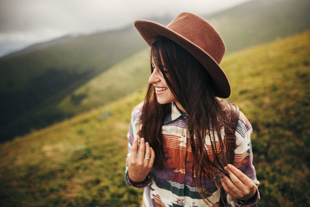 stylish traveler hipster girl in hat, with backpack and windy hair, smiling in mountains in clouds. space for text. amazing atmospheric moment. travel and wanderlust conceptの写真素材
