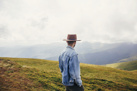 stylish traveler man in hat standing on top of sunny mountains in clouds. space for text. hipster...