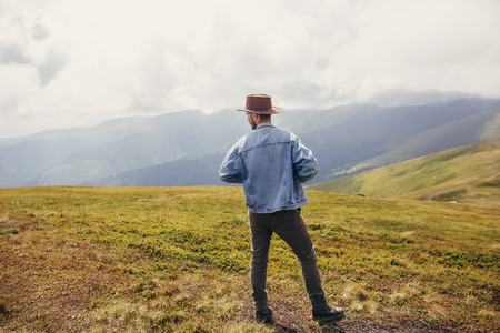 travel and wanderlust concept. stylish traveler man in hat standing on top of sunny mountains in...
