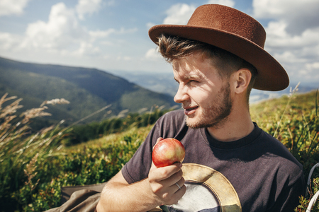 stylish traveler man in hat eating apple on top of sunny mountains. travel and wanderlust concept. space for text. happy hipster traveling and camping. summer vacationの写真素材