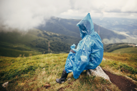 travel and wanderlust concept. traveler hipster girl in blue raincoat with backpack, exploring misty mountains. space for text. atmospheric moment.  woman sitting on top hillの写真素材
