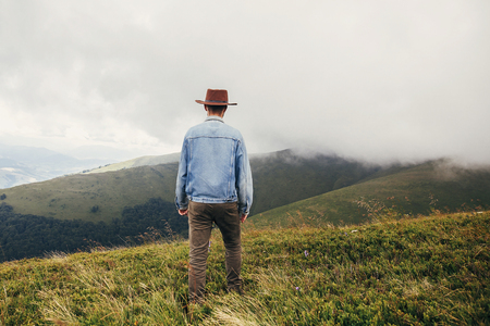 stylish traveler man in hat walking on top of sunny mountains into the  misty clouds. travel and wanderlust concept. space for text. happy hipster guy traveling. summer vacationの写真素材