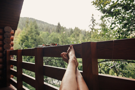 girl relaxing in wooden cottage on porch among woods. woman legs on balcony with view on forest in mountains. atmospheric moment. summer vacationの写真素材