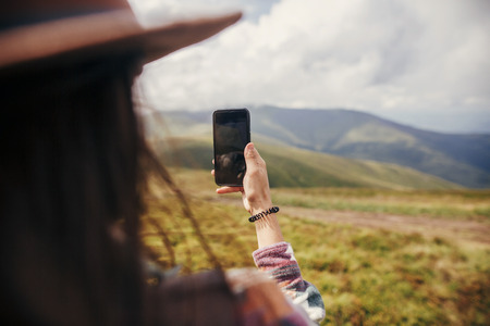 stylish traveler girl in hat with windy hair holding phone on top of sunny mountains. summer vacation. travel and wanderlust concept. space for text. back view. amazing atmospheric momentの写真素材