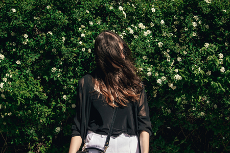 beautiful Stylish Hipster Girl with beautiful windy hair having fun at floral bushes in sunny day. Boho Woman with sunglasses in fashionable outfit, smiling and enjoying day in gardenの写真素材