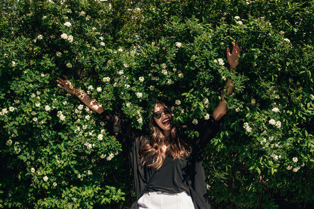 beautiful Stylish Hipster Girl with beautiful hair relaxing at floral bushes in sunny day. Happy Boho Woman with sunglasses in fashionable outfit enjoying day in flowers in gardenの写真素材