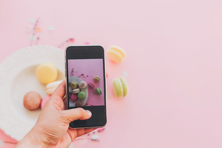 blogging concept, flat lay. food photography. hand holding phone and taking photo of stylish colorful macaroons in vintage plate on trendy pink paper. space for textの写真素材