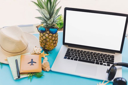 planning summer vacation concept. stylish laptop with empty screen, pineapple in sunglasses and passport, map, hat, shells, notebook on trendy blue paper table. travel and wanderlustの写真素材