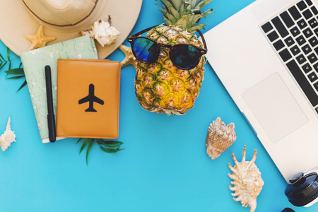 planning summer vacation concept. stylish laptop, passport, pineapple in sunglasses, map, hat, headphones, shells, notebook on trendy blue paper. flat lay. travel and wanderlustの写真素材