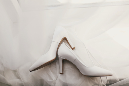 beautiful stylish shoes on luxury silk wedding dress, flat lay. simple white shoes on white gown with lace floral ornaments, bridal morning preparations. top viewの写真素材