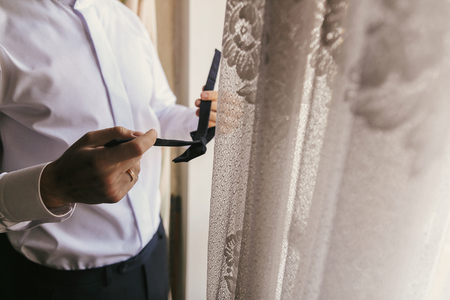 groom in white shirt holding bow tie and getting ready near window. morning preparations before wedding day. groom outfit.の写真素材