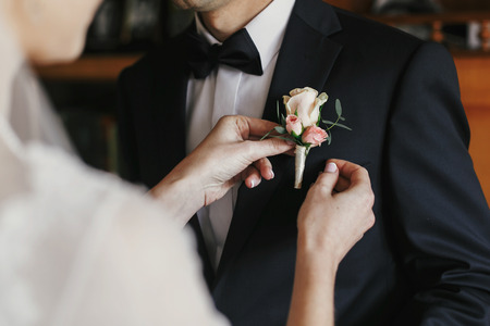 beautiful bride putting on stylish simple boutonniere with roses on groom black suit. wedding morning preparationsの写真素材