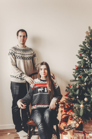 stylish hipster family couple in stylish sweaters sitting under christmas tree with lights and posing. merry christmas and happy new year concept. space for textの写真素材