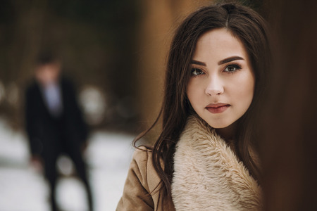 gorgeous bride in coat looking and stylish groom walking under trees in winter forest. happy wedding couple posingg in winter snowy park. romantic sensual moment of newlywedsの写真素材
