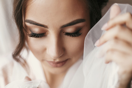 gorgeous bride portrait with make-up in the morning in room near window. beautiful woman getting ready for wedding day, holding veil and smiling. face close up. sensual momentの写真素材