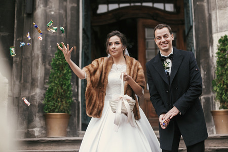 gorgeous bride in coat and stylish groom throwing candy near church door after wedding ceremony.  happy newlywed couple smiling and having fun. romantic  momentの写真素材