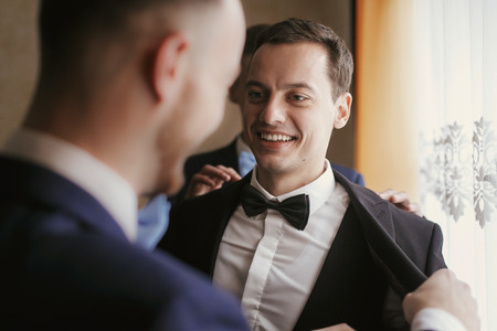 Groom getting ready in the morning with groomsmen in the room. man hands putting on jacket and bow tie, standing at window light in hotel. preparation for wedding dayの写真素材