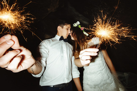 happy beautiful bride and elegant stylish groom holding fireworks on the top of a mountains in the eveningの写真素材