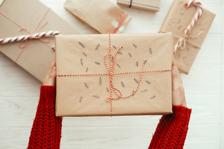 hands holding stylish christmas present with red ribbon and tree pattern. flat lay. modern craft gifts on white wooden background. seasonal greetings. merry christmas conceptの写真素材