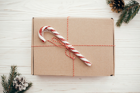 stylish simple christmas present with red ribbon, candy cane, pine branches and cones. modern craft gift on white wooden background, flat lay. seasonal greetings card.の写真素材