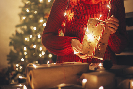 merry christmas concept. hands holding christmas present in lights in evening festive room under tree in illumination. space for text. girl opening modern craft gifts with candy caneの写真素材